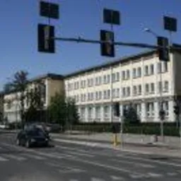 Ulica miejska z przejściem dla pieszych i sygnalizacją świetlną na tle dużego, dwupiętrowego budynku z licznymi oknami. Czarny samochód osobowy przejeżdża przez skrzyżowanie.