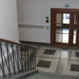 Klatka schodowa w budynku biurowym z drewnianą balustradą i drzwiami. Widoczne ściany pomalowane na biało z szarym pasem na wysokości poręczy, marmurowa podłoga w szaro-beżową kratę...