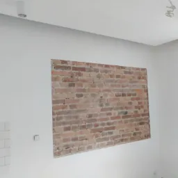 Fragment ściany z odsłoniętą czerwoną cegłą, otoczony świeżo pomalowaną, białą ścianą. Obok fragment ściany z białymi, prostokątnymi płytkami. Widoczny włącznik elektryczny.