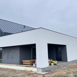 Domy i Domki Drewniane - Nowoczesny dom szkieletowy w trakcie budowy: białe ściany, grafitowy dach z blachy na rąbek stojący, duże okna, widoczne materiały budowlane i teren wokół.