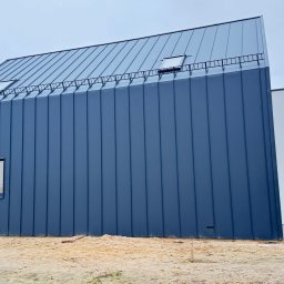 Domy i Domki Drewniane - Nowoczesny dom szkieletowy z ciemną blachą na elewacji i dachu, widok z boku. Małe okno na ścianie, drabinka na dachu. Teren wokół w trakcie prac wykończeniowych.