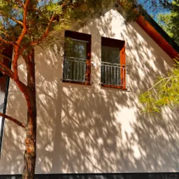 Biały dom szkieletowy z balkonami, otoczony drzewami iglastymi, w słoneczny dzień. Cień drzew na elewacji. Widok z zewnątrz, podkreślający naturalne otoczenie.