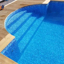 Nowoczesny basen z niebieską mozaiką i drewnianym wykończeniem tarasu, widoczne schody w basenie, na drewnianym tarasie leżą narzędzia i akcesoria.