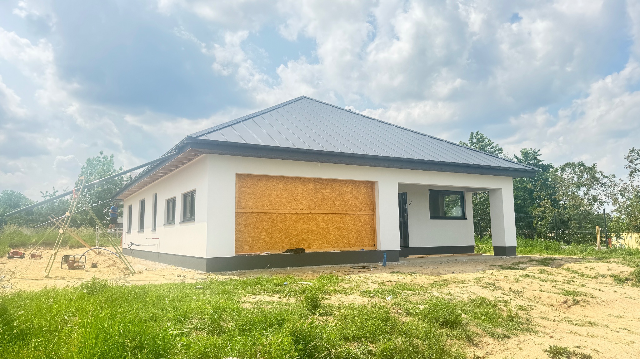 Biały dom szkieletowy z szarą blachodachówką w trakcie budowy. Widoczne rusztowania i płyty OSB zamiast bramy garażowej. Letni dzień, zielona trawa.