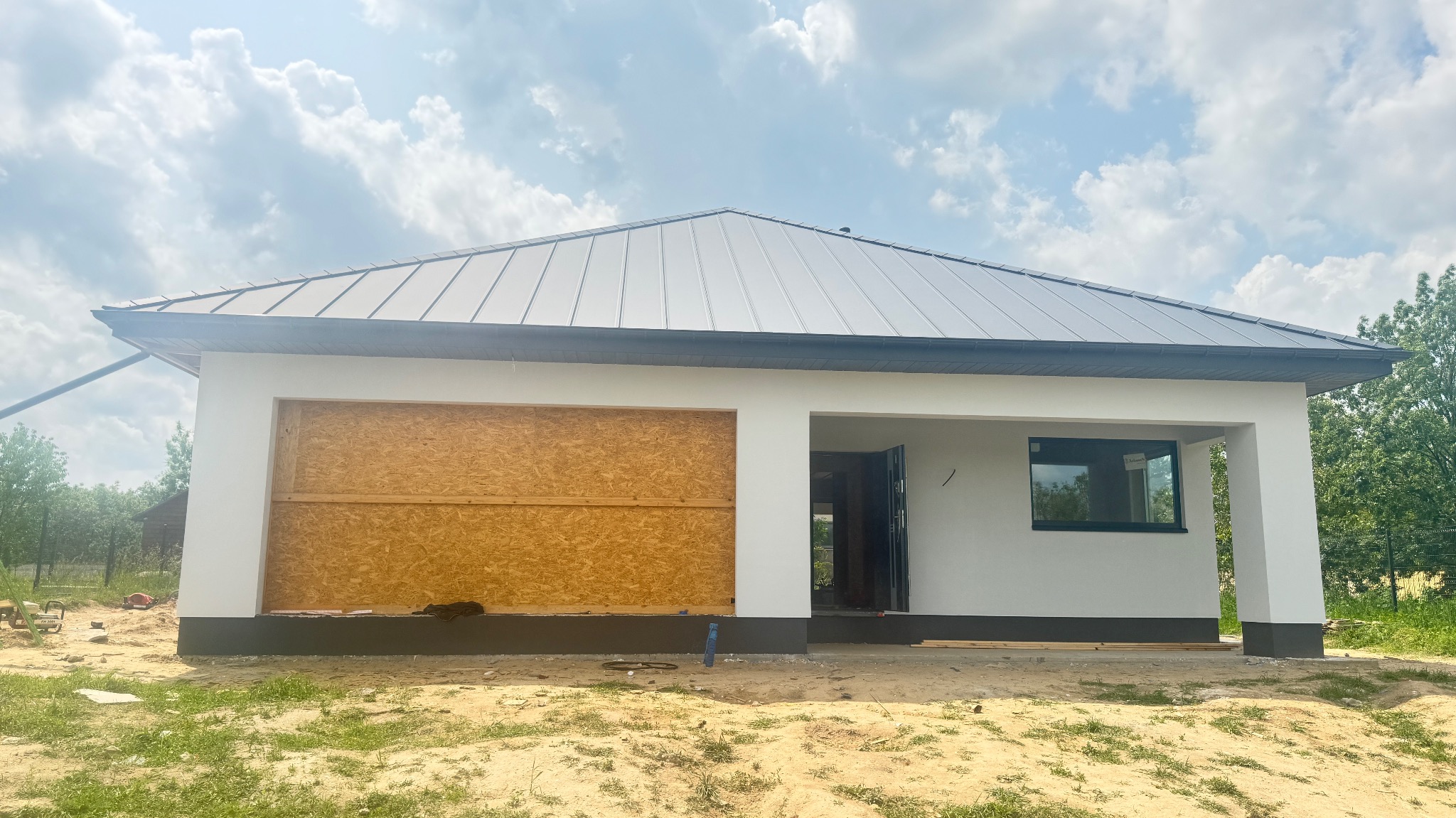 Nowoczesny, parterowy dom w trakcie budowy: białe ściany, szary dach, okno z widokiem na zieleń. Jedna ściana zabezpieczona płytą OSB. Ujęcie w słoneczny dzień.