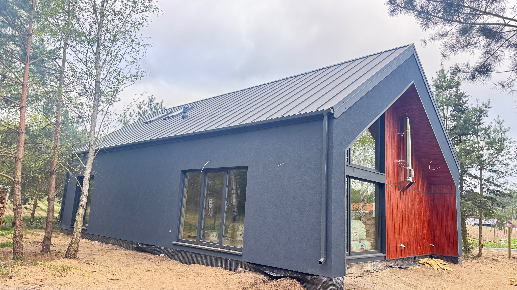 Nowoczesny dom szkieletowy z ciemną elewacją i drewnianym szczytem, widok z zewnątrz na tle drzew i pochmurnego nieba. Widoczne okna i komin.