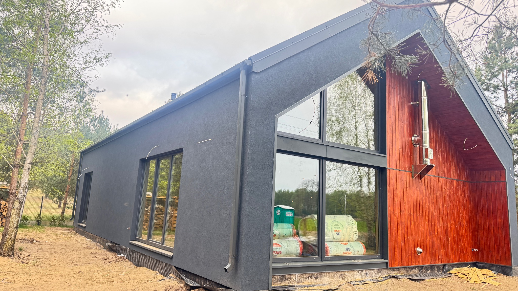 Nowoczesny dom szkieletowy z dużymi oknami i kominem na drewnianej elewacji, otoczony zielenią. Wewnątrz widoczne rolki materiału izolacyjnego.