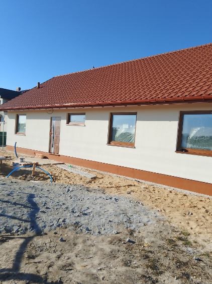 Gotowy dom szkieletowy z czerwoną dachówką i oknami zabezpieczonymi folią. Widoczna elewacja w jasnym kolorze i fragment terenu przed budynkiem. Słoneczny dzień.