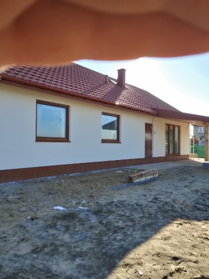 Jasny dom z brązowym dachem z blachodachówki, brązowymi oknami i podbitką. Widoczny fragment dłoni w kadrze. Teren wokół domu niezagospodarowany.