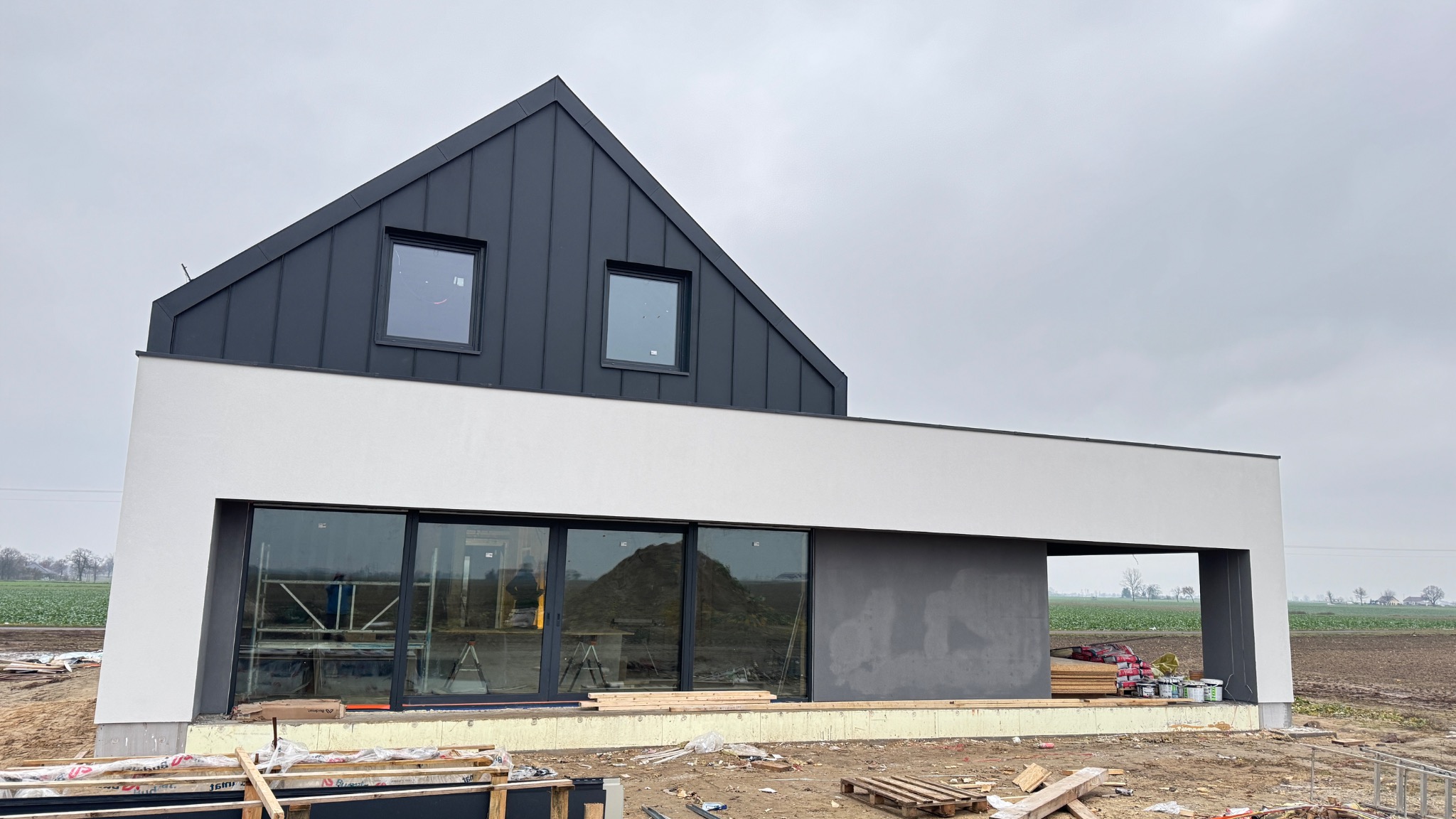 Nowoczesny dom w trakcie budowy z czarnym dachem, białymi ścianami i dużymi oknami, otoczony polami. Widoczne materiały budowlane i rusztowania.