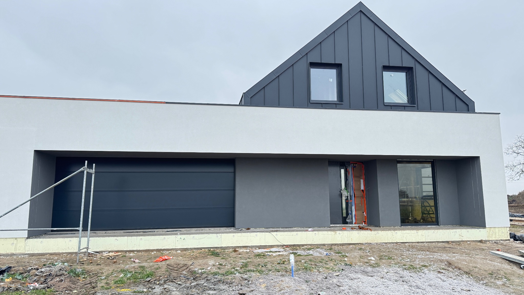 Nowoczesny dom z szarą blachą na dachu i białymi ścianami, widoczny garaż z bramą segmentową. Na pierwszym planie teren budowy z elementami izolacji.