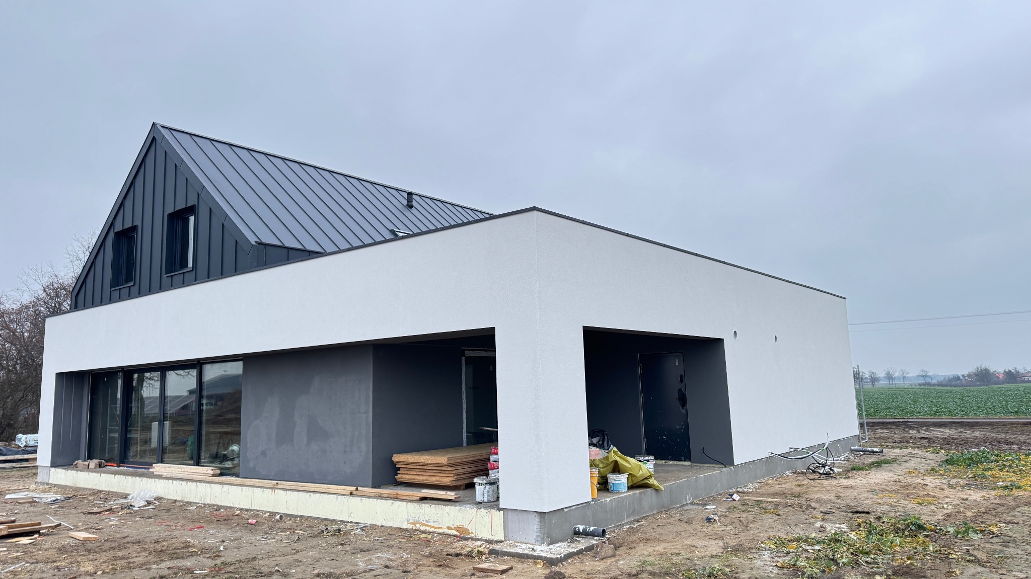 Nowoczesny dom szkieletowy w trakcie budowy: białe ściany, grafitowy dach z blachy na rąbek stojący, duże okna, widoczne materiały budowlane i teren wokół.