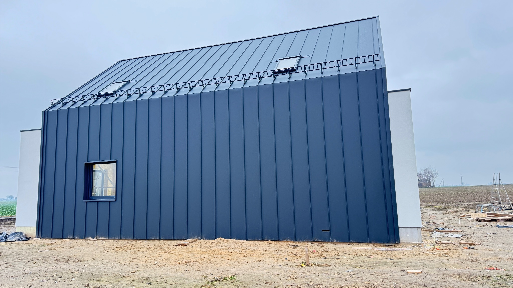 Nowoczesny dom szkieletowy z ciemną blachą na elewacji i dachu, widok z boku. Małe okno na ścianie, drabinka na dachu. Teren wokół w trakcie prac wykończeniowych.