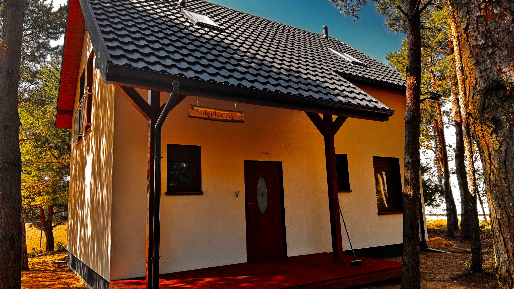 Gotowy, parterowy dom szkieletowy z ciemną dachówką, drewnianą werandą i tabliczką adresową 'Nadwiślańska 47', otoczony drzewami w słoneczny dzień.
