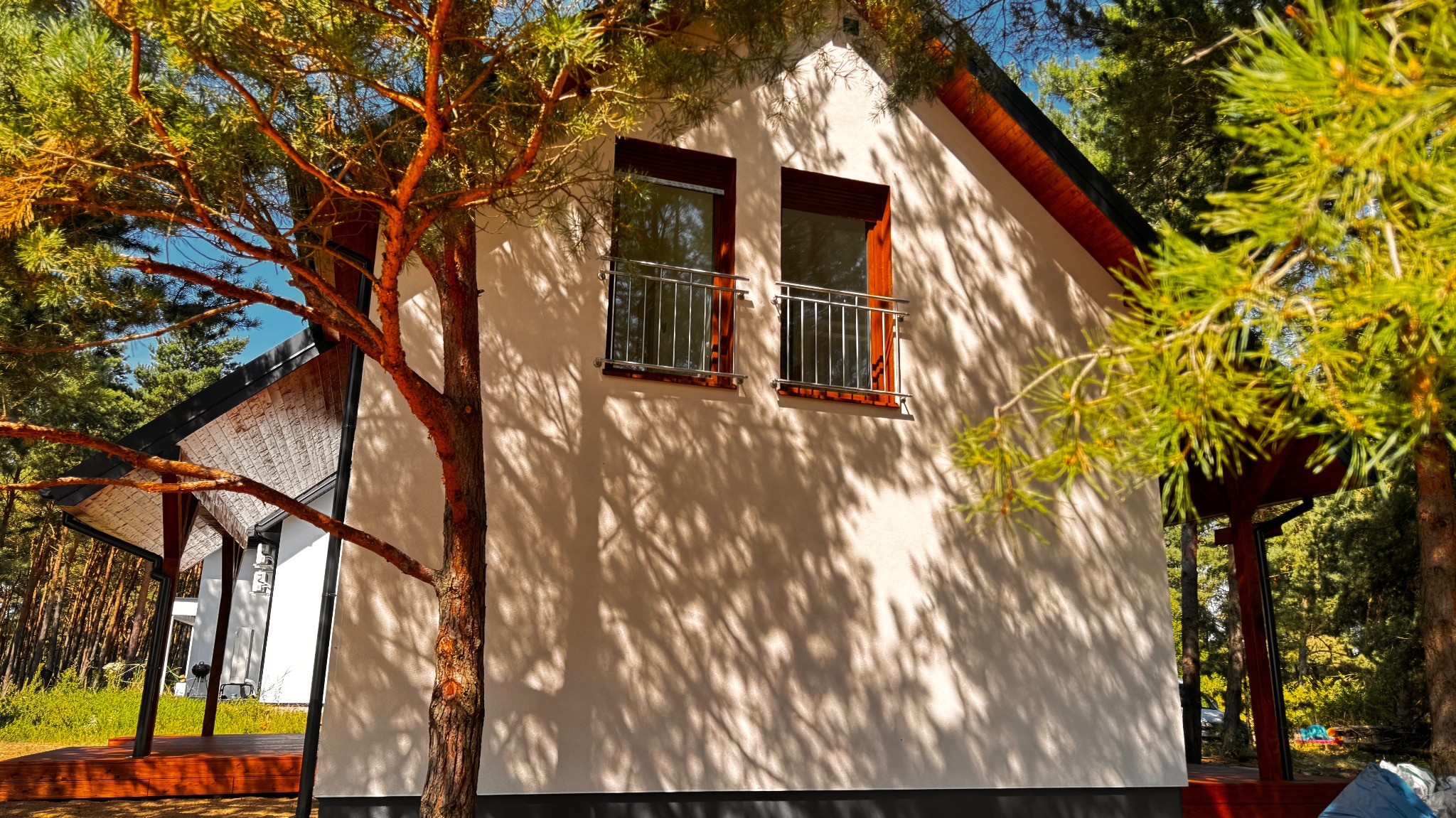 Biały dom szkieletowy z balkonami, otoczony drzewami iglastymi, w słoneczny dzień. Cień drzew na elewacji. Widok z zewnątrz, podkreślający naturalne otoczenie.