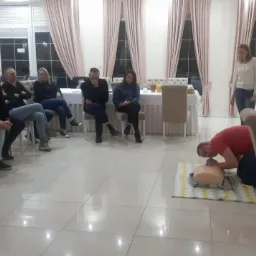 Instruktor demonstruje resuscytację krążeniowo-oddechową na manekinie przed grupą kursantów w jasnym pomieszczeniu z oknami i zasłonami.