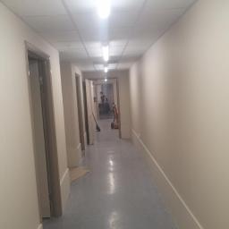 uslugi remontowe ADRIAN - Długi korytarz po remoncie, świeżo pomalowane ściany w odcieniach beżu, szara podłoga, widoczne drzwi i osoba w oddali, sprzęt do sprzątania na końcu korytarza.