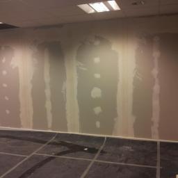 uslugi remontowe ADRIAN - Ściana w trakcie szpachlowania z widocznymi paskami świeżo nałożonej masy szpachlowej, ciemna podłoga zabezpieczona folią, widoczne otwory po gniazdkach elektrycznych.