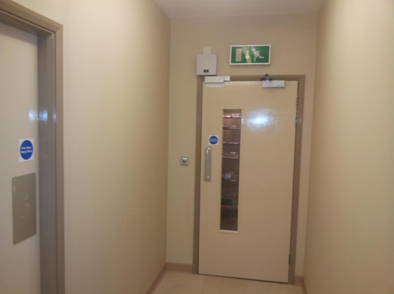 Korytarz z dwiema parami drzwi przeciwpożarowych w beżowych odcieniach, jedna z podłużnym okienkiem, oznakowanie ewakuacyjne i gaśnicze na ścianach.