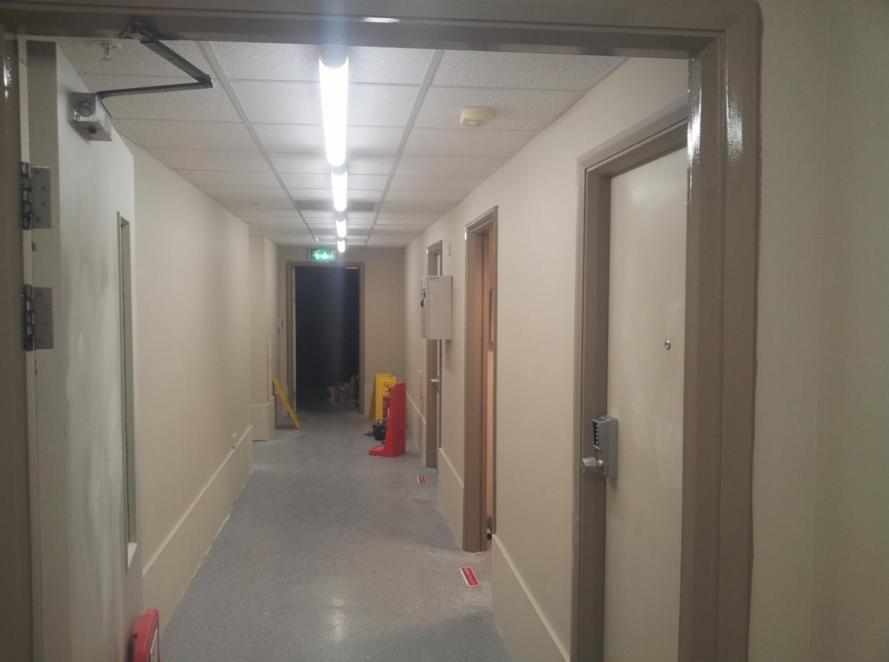 Długi korytarz w trakcie odświeżania, widoczne świeżo pomalowane ściany w odcieniach beżu, zabezpieczenia podłogi i drzwi, w głębi korytarza sprzęt malarski i tablica ostrzegawcza.