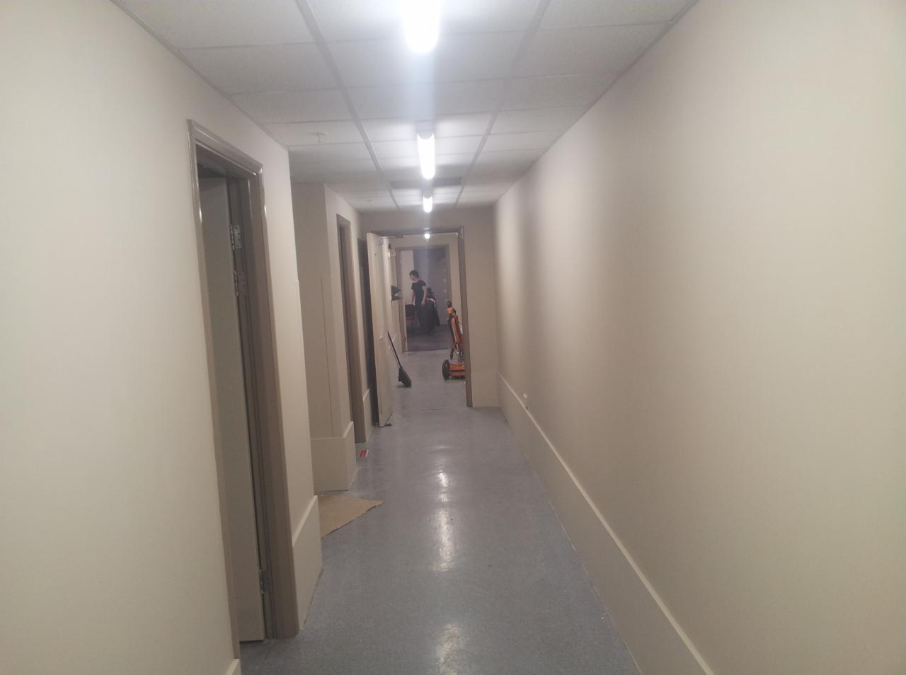 Długi korytarz po remoncie, świeżo pomalowane ściany w odcieniach beżu, szara podłoga, widoczne drzwi i osoba w oddali, sprzęt do sprzątania na końcu korytarza.