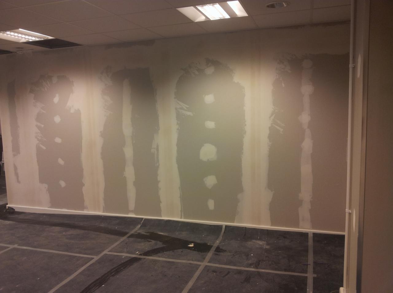 Ściana w trakcie szpachlowania z widocznymi paskami świeżo nałożonej masy szpachlowej, ciemna podłoga zabezpieczona folią, widoczne otwory po gniazdkach elektrycznych.