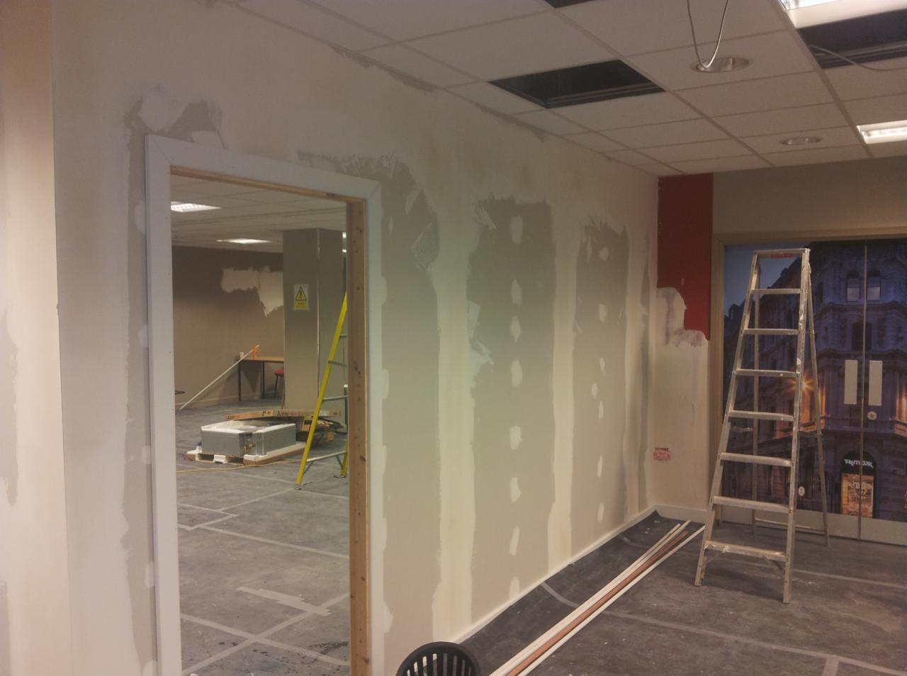 Wnętrze w trakcie prac wykończeniowych: ściany z nałożoną gładzią, widoczne ślady po łatach, drabina oparta o ścianę z fototapetą, otwór drzwiowy z ościeżnicą w trakcie obróbki, żółta drabina w tle.