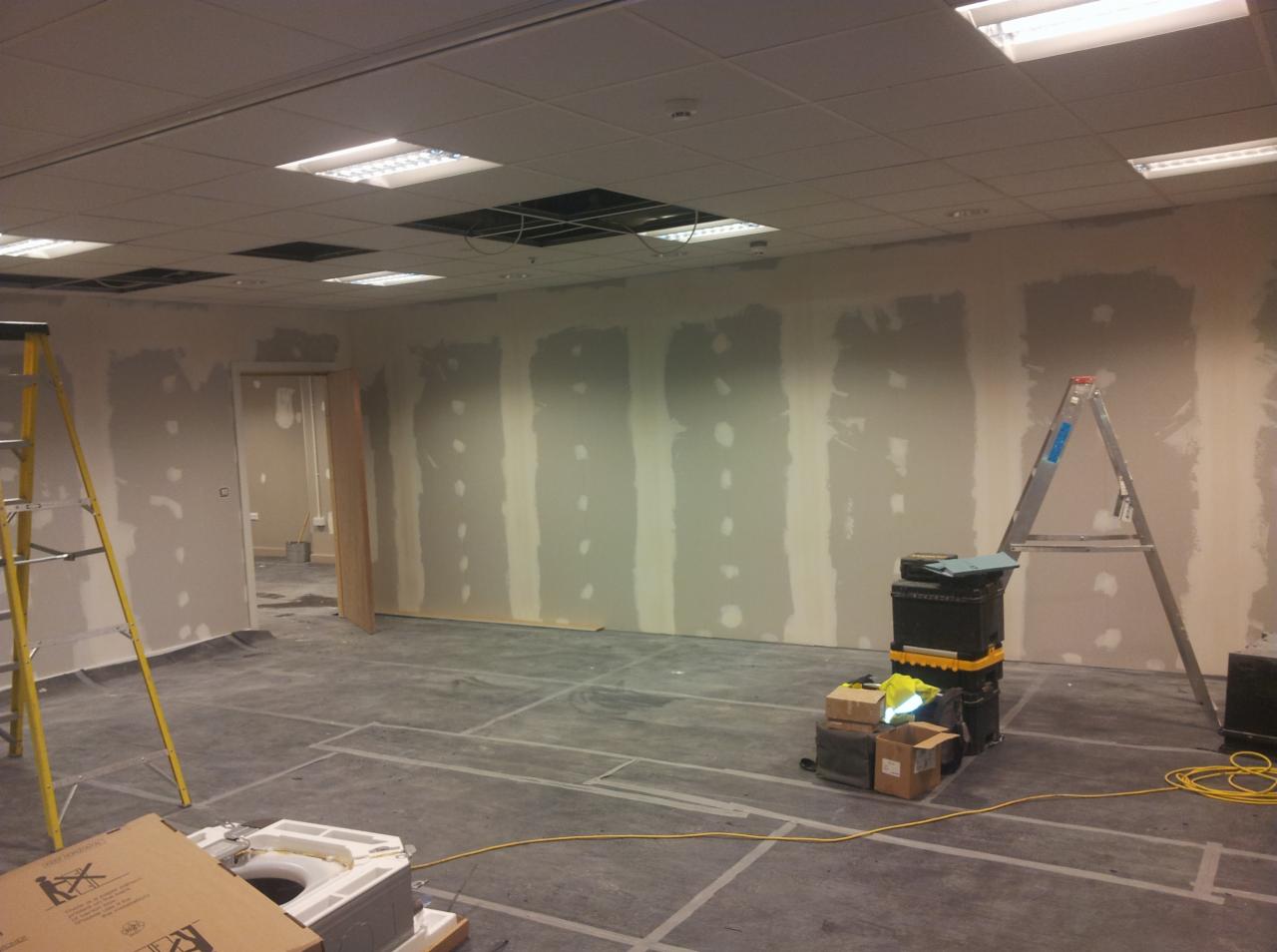 Wnętrze biurowe w trakcie wykańczania: ściany z widocznymi miejscami po szpachlowaniu, podwieszany sufit z otworami, drabiny, narzędzia i materiały budowlane na podłodze.