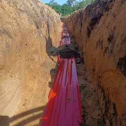 Energomontaż mgr inż. Zdzisław Adamczyk - Wykop w ziemi z ułożoną czerwoną folią zabezpieczającą, prawdopodobnie nad instalacją elektryczną. Widoczny fragment łopaty i dwóch osób w oddali.