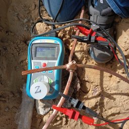 Energomontaż mgr inż. Zdzisław Adamczyk - Miernik Metrel podłączony klemami do miedzianych przewodów uziemiających w piasku, pomiar rezystancji uziemienia, widoczne buty osoby nadzorującej pomiar.