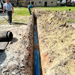 Energomontaż mgr inż. Zdzisław Adamczyk - Wykop z rurami w niebieskiej folii, widoczni dwaj mężczyźni przy pracy. Na drugim planie budynek gospodarczy i sprzęt budowlany.