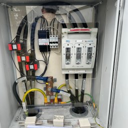 Energomontaż mgr inż. Zdzisław Adamczyk - Otwarta szafa elektryczna z bezpiecznikami, okablowaniem oznaczonym etykietami, elementami miedzianymi i dokumentacją techniczną na dnie. Widoczne elementy montażowe.