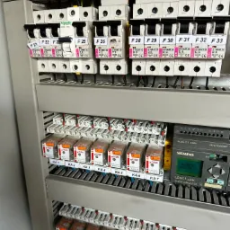 Szczegółowe ujęcie rozdzielnicy elektrycznej z bezpiecznikami, przekaźnikami i modułem sterującym Siemens LOGO!, ukazujące złożoność instalacji.