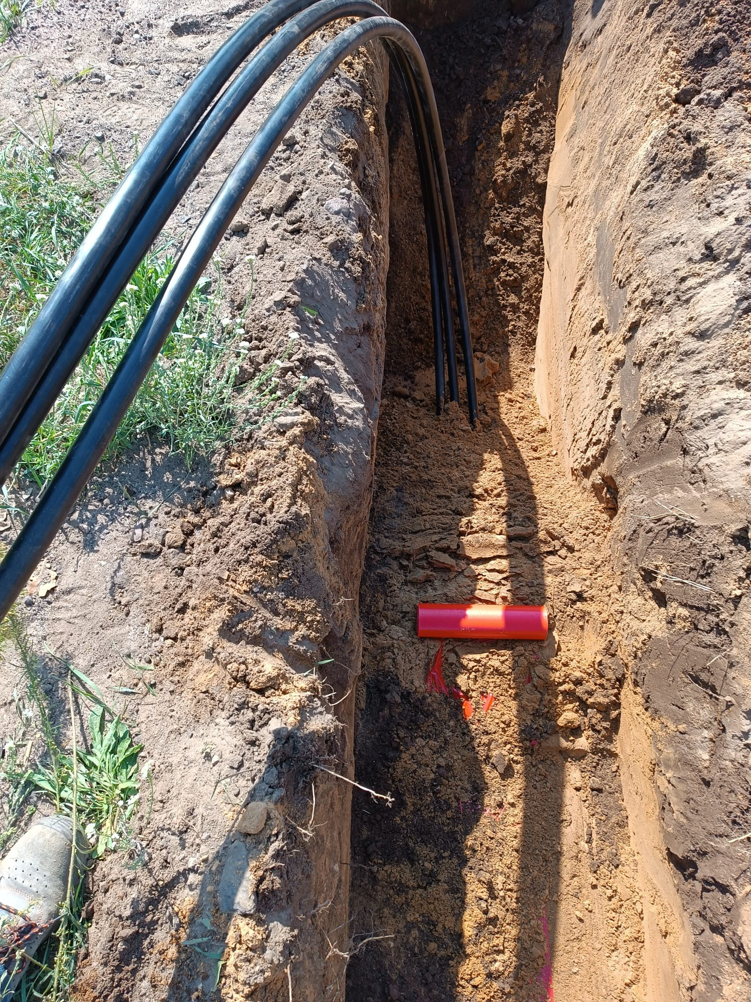 Wykop z ułożonymi kablami w czarnej izolacji oraz pomarańczową rurą osłonową. Widoczna ziemia i trawa. Kable ułożone równolegle.