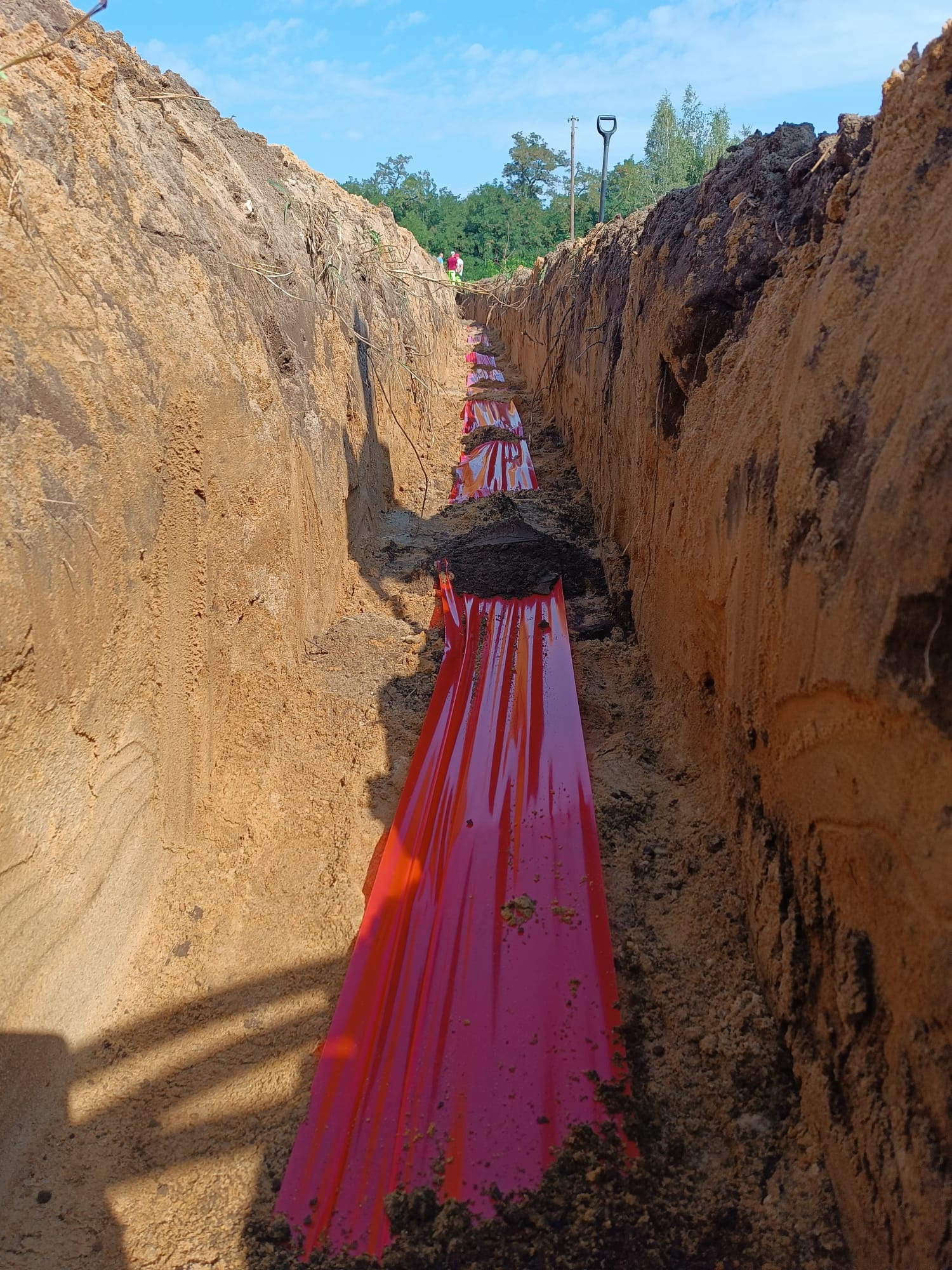 Wykop w ziemi z ułożoną czerwoną folią zabezpieczającą, prawdopodobnie nad instalacją elektryczną. Widoczny fragment łopaty i dwóch osób w oddali.
