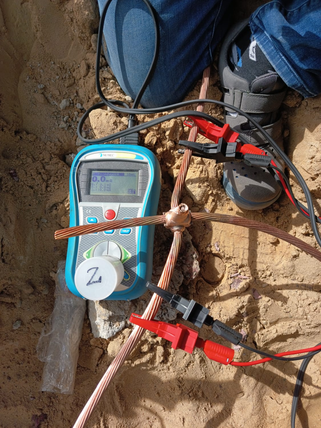 Miernik Metrel podłączony klemami do miedzianych przewodów uziemiających w piasku, pomiar rezystancji uziemienia, widoczne buty osoby nadzorującej pomiar.