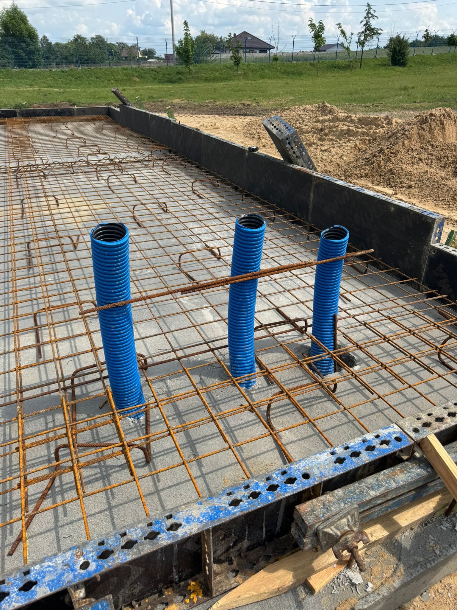 Fundament zbrojony z niebieskimi rurami przepustowymi, przygotowany do zalania betonem. Widok z góry, w tle zieleń i błękitne niebo.