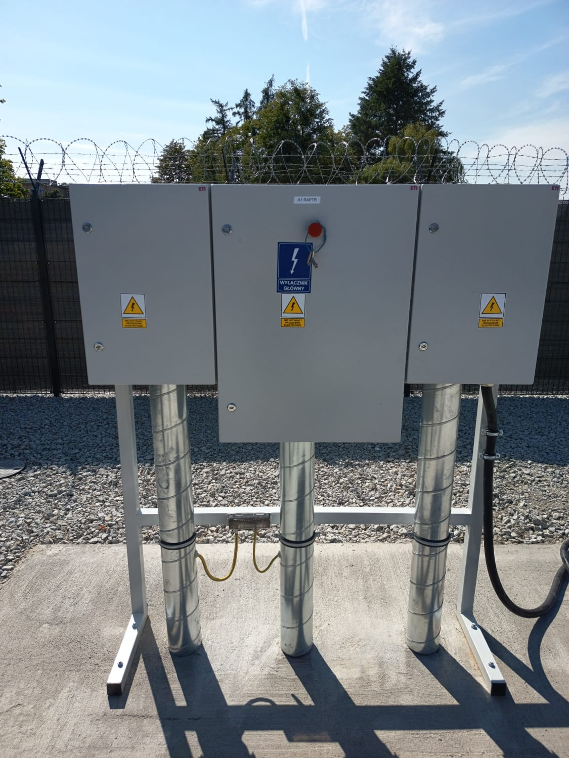 Szare szafy elektryczne z żółtymi oznaczeniami ostrzegawczymi i wyłącznikiem głównym, umieszczone na metalowych rurach, otoczone drutem kolczastym na tle nieba.