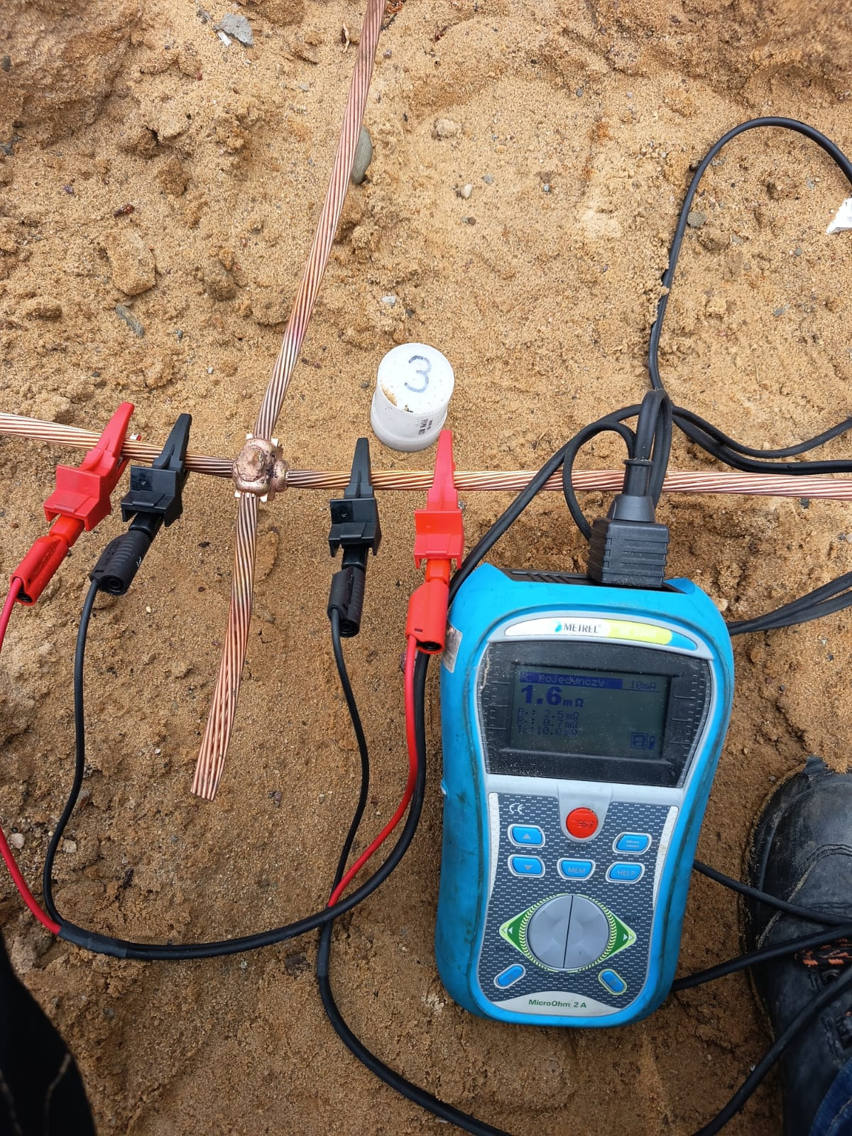 Miernik podłączony do uziemienia w piasku, pomiar rezystancji uziomu. Widoczne kable, krokodylki pomiarowe i wyświetlacz urządzenia.