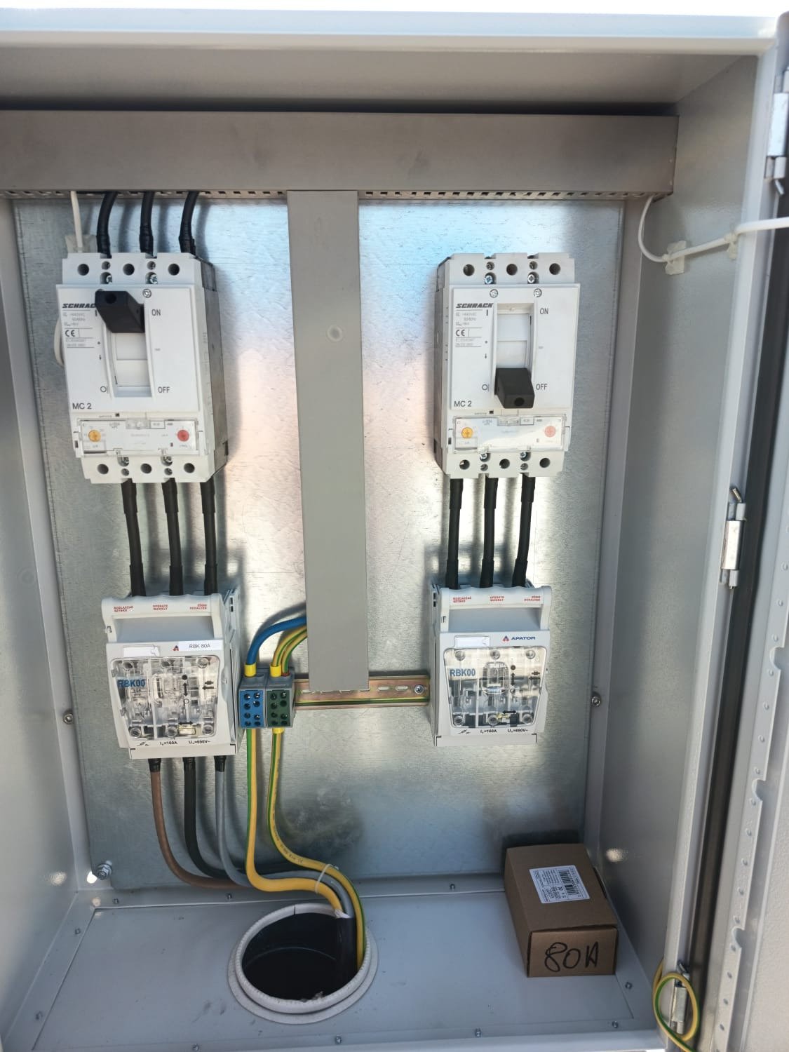 Wnętrze metalowej skrzynki z zamontowanymi dwoma licznikami i wyłącznikami. Widoczne przewody, listwa zaciskowa i otwór na kable. Skrzynka z osprzętem elektrycznym.