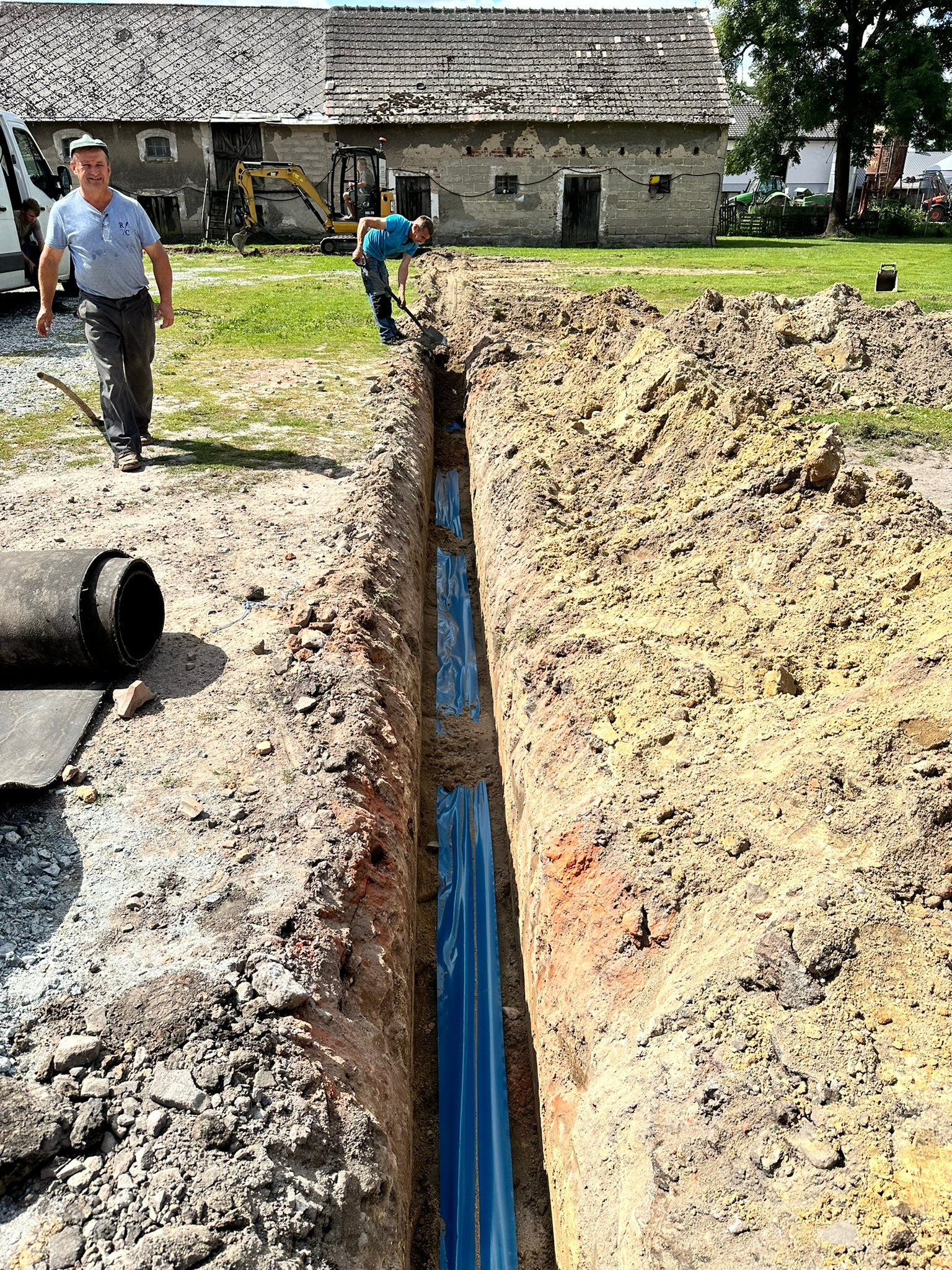 Wykop z rurami w niebieskiej folii, widoczni dwaj mężczyźni przy pracy. Na drugim planie budynek gospodarczy i sprzęt budowlany.