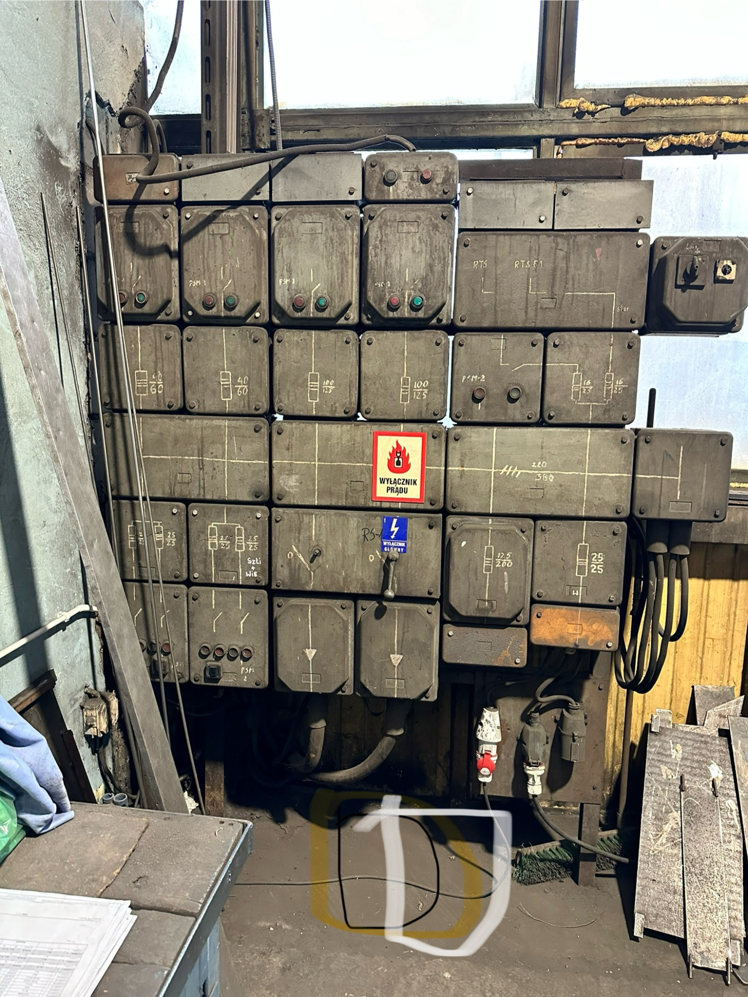 Rozdzielnia elektryczna z wyłącznikami i oznaczeniami, widoczne przewody, tabliczki ostrzegawcze. Szara, przemysłowa instalacja elektryczna.