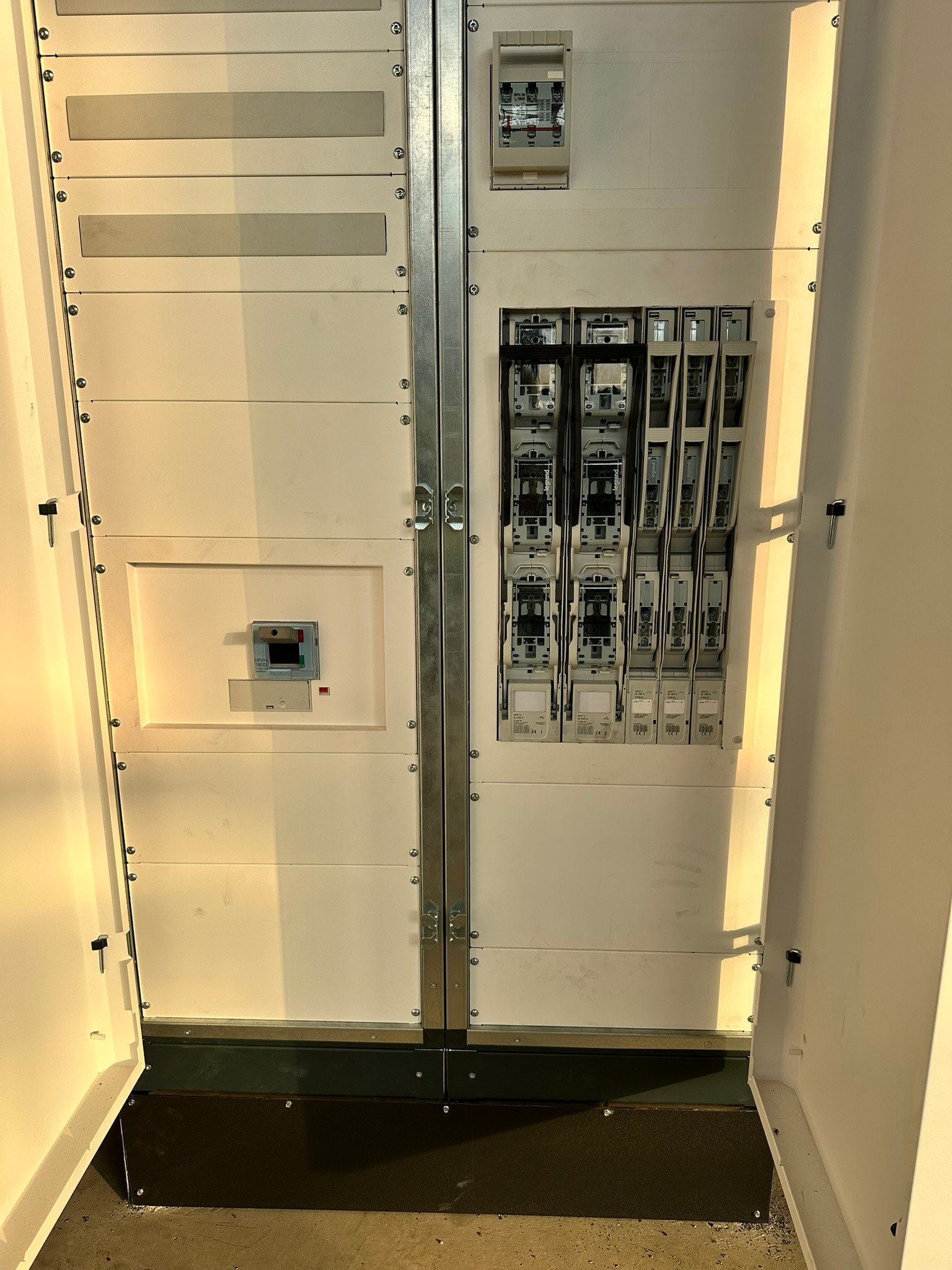 Otwarta, biała szafa elektryczna z widocznymi bezpiecznikami i elementami sterującymi, oświetlona z lewej strony, ukazująca wnętrze rozdzielnicy.