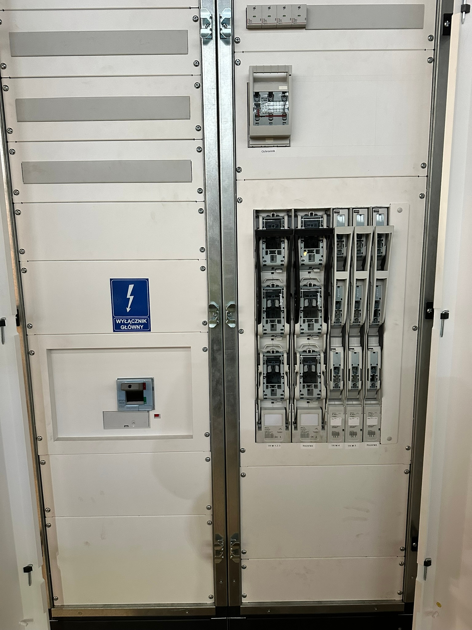 Otwarta szafa elektryczna z bezpiecznikami, wyłącznikiem głównym i modułami rezerwowymi. Widoczne szczegóły montażu i okablowania wewnątrz.