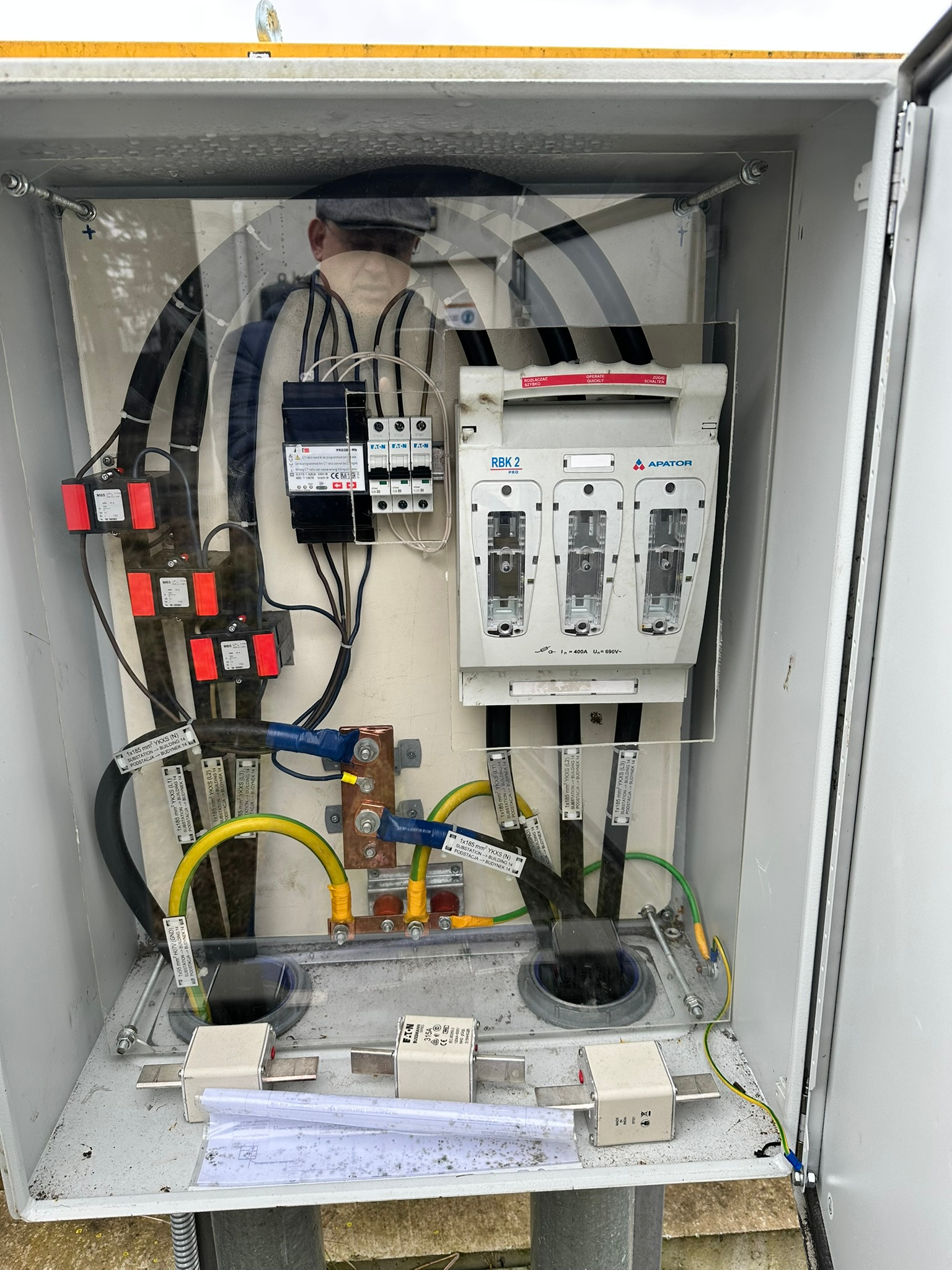 Otwarta szafa elektryczna z bezpiecznikami, okablowaniem oznaczonym etykietami, elementami miedzianymi i dokumentacją techniczną na dnie. Widoczne elementy montażowe.