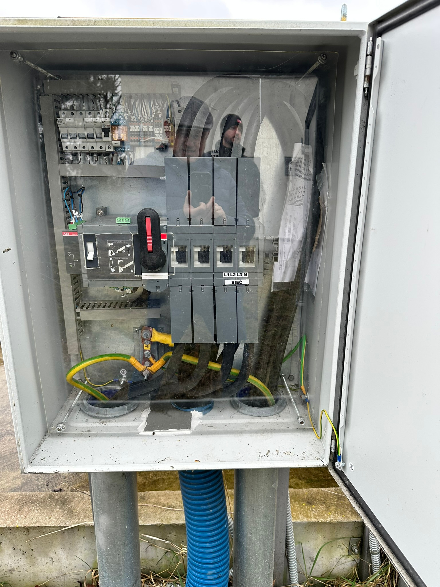 Otwarta skrzynka elektryczna z widocznymi bezpiecznikami, przewodami i elementami przyłączeniowymi. Refleksja osoby widoczna na przezroczystej osłonie.