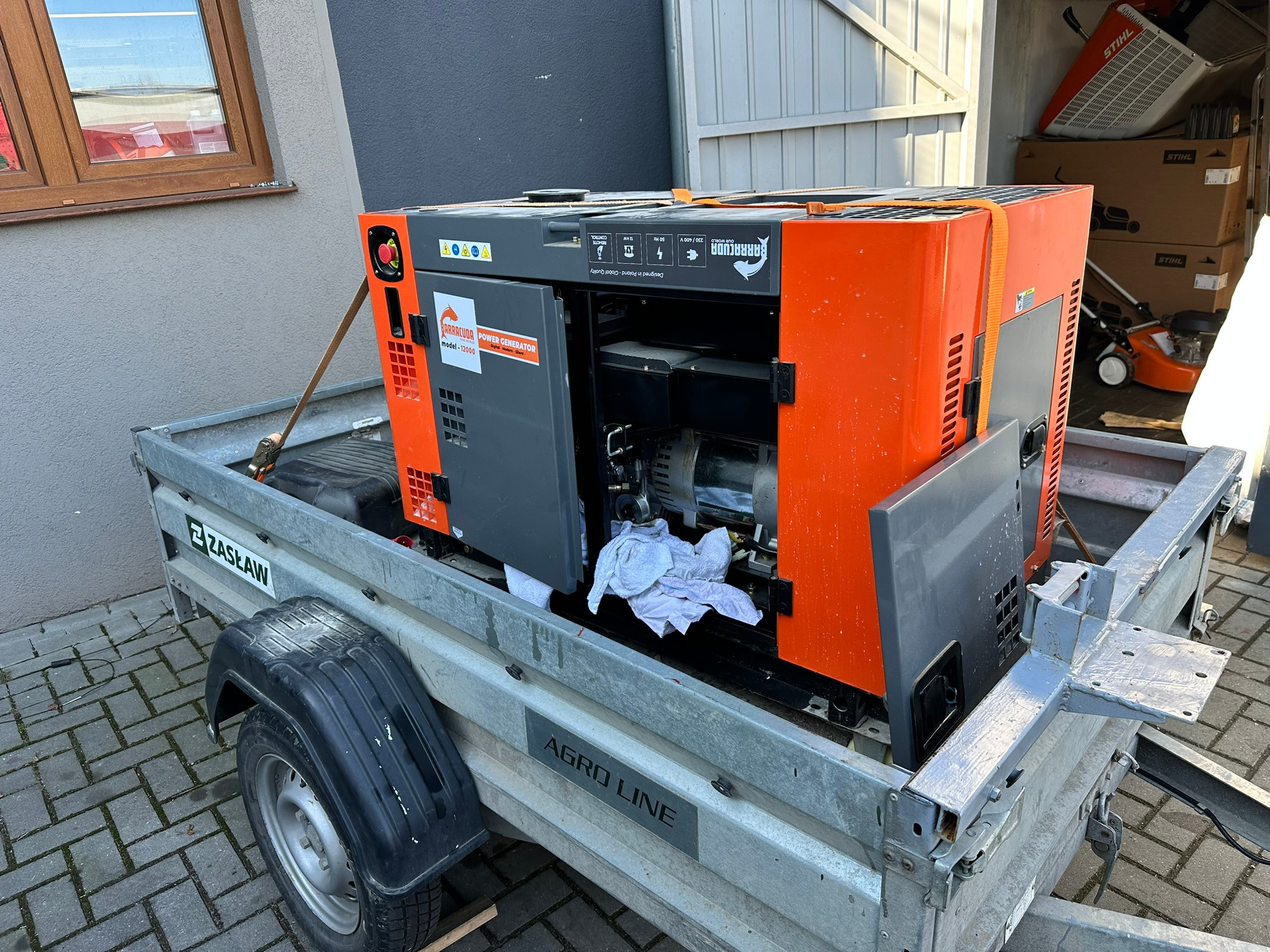 Agregat prądotwórczy Barracuda model I2000 na przyczepie ZASŁAW, z otwartą obudową, widoczne wnętrze silnika. W tle skrzynie i kosiarka STIHL.