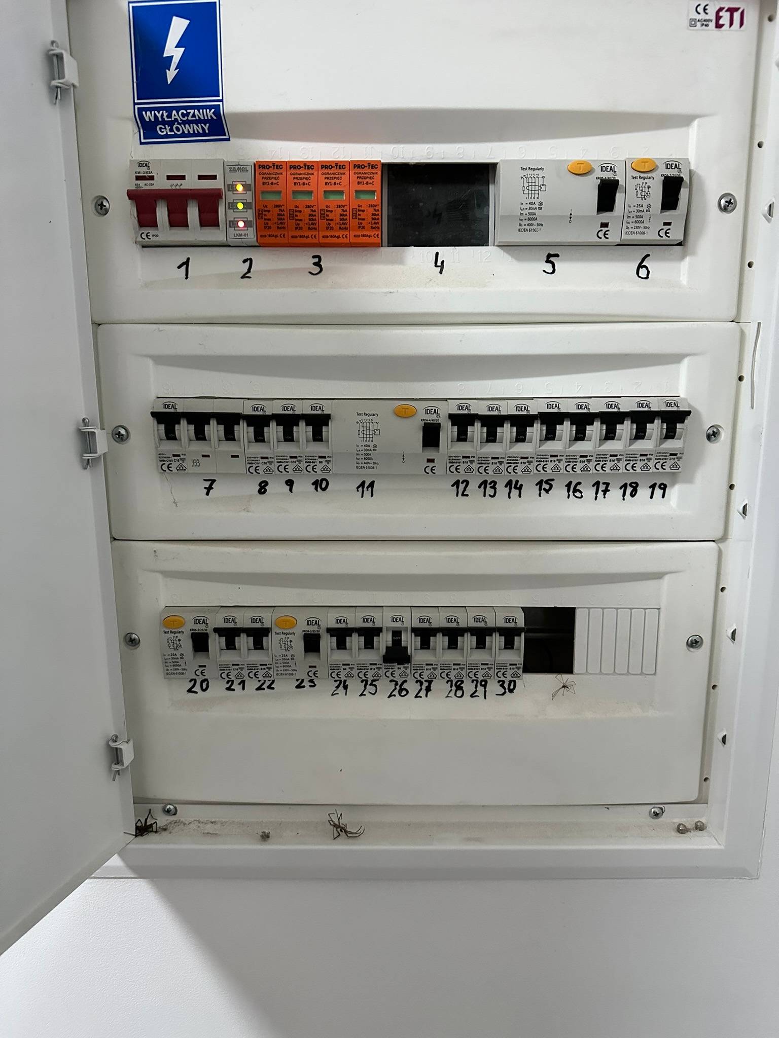 Otwarta skrzynka elektryczna z bezpiecznikami i wyłącznikami, widoczne oznaczenia i numery na białej obudowie. Niebieska naklejka z symbolem błyskawicy.