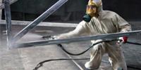Pracownik w kombinezonie ochronnym i masce maluje natryskowo metalową konstrukcję hali na szaro.