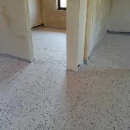 Szara, nakrapiana posadzka w surowym wnętrzu, widok na dwa pomieszczenia z widocznymi niedoskonałościami ścian i nielicznymi plamami.